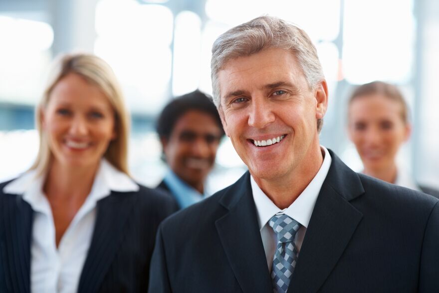 A grey-haired principal smiling, with colleagues out-of-focus behind.