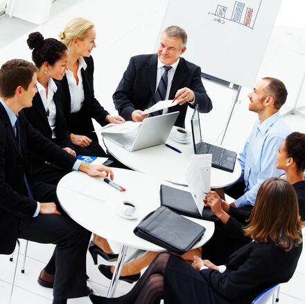 A senior director leads a real meeting around a circular table with five colleagues.